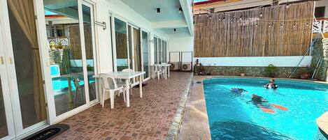 A swimming pool with a tiled deck, a covered patio area with a table and chairs, and a wooden fence in the background.