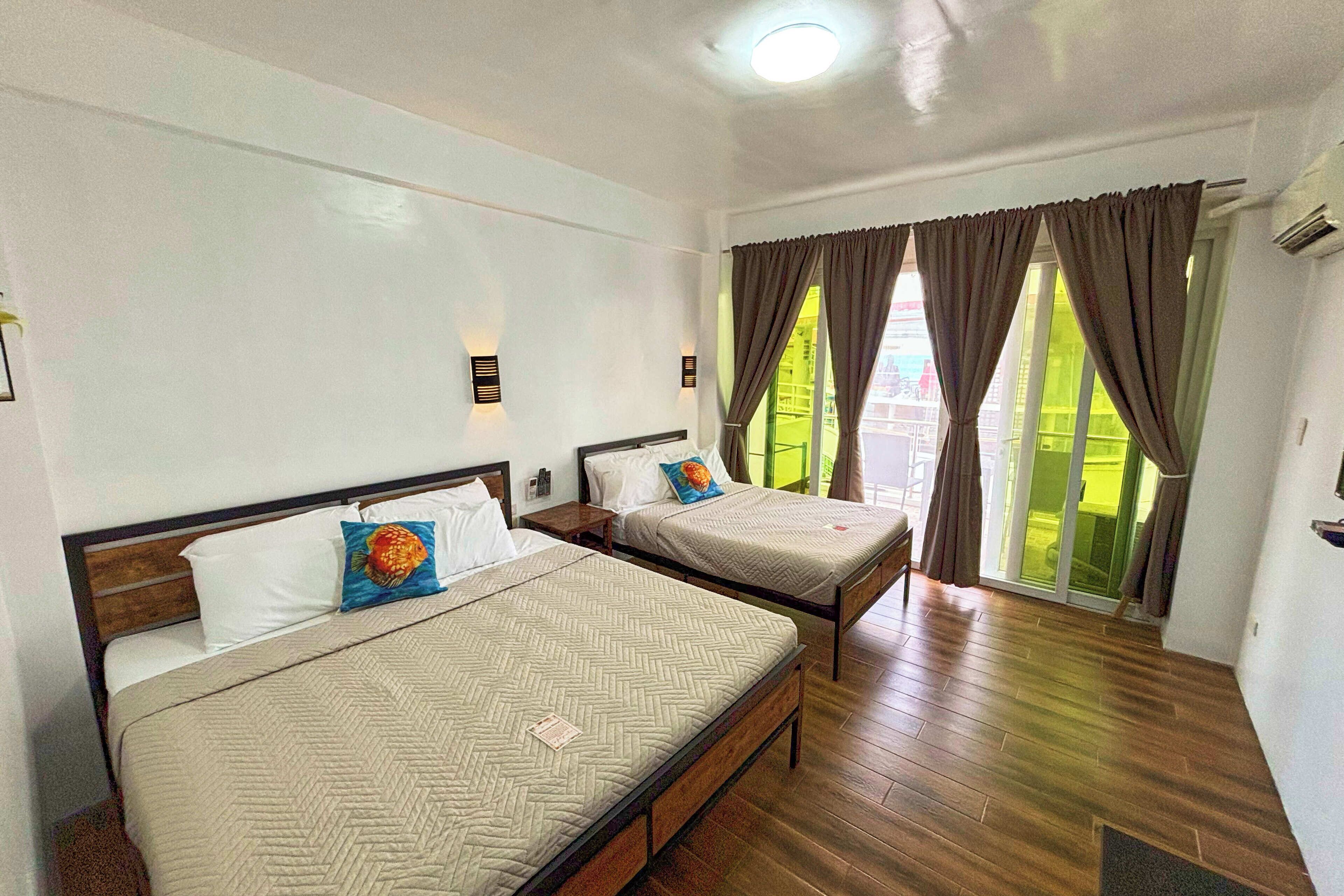 A hotel room with two beds, wooden flooring, and a view of a green outdoor area through the window.
