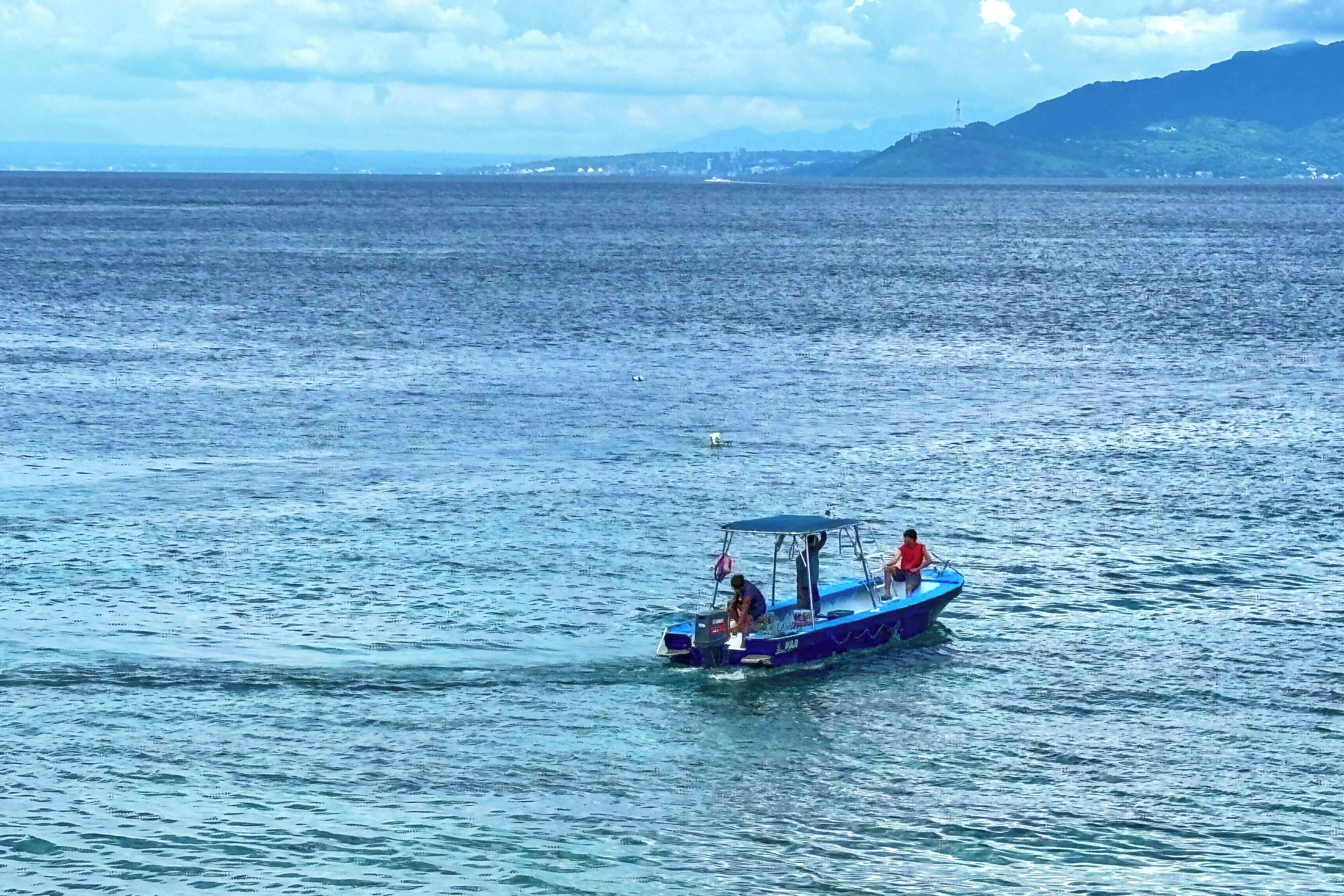 A boat with a canopy and two people on it, moving on a calm sea with distant land and mountains.