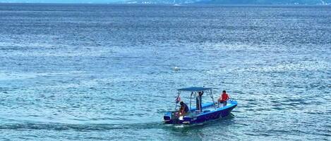 A boat with a canopy and two people on it, moving on a calm sea with distant land and mountains.