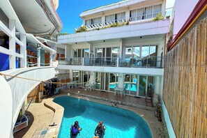 Uma área de piscina em vários níveis, com duas pessoas em equipamento de mergulho, um deck coberto e um edifício com varandas e janelas.