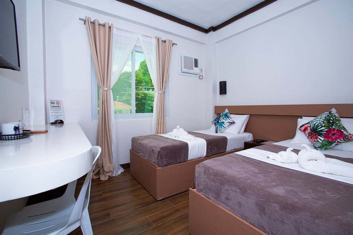 A hotel room with two beds, a desk, a chair, a window with curtains, and a TV.