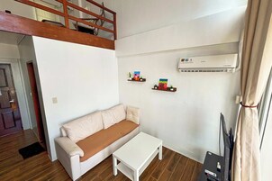 Una sala de estar con un sofá, una mesa de centro y un televisor. Hay un altillo de madera sobre el sofá, y un equipo de aire acondicionado en la pared.