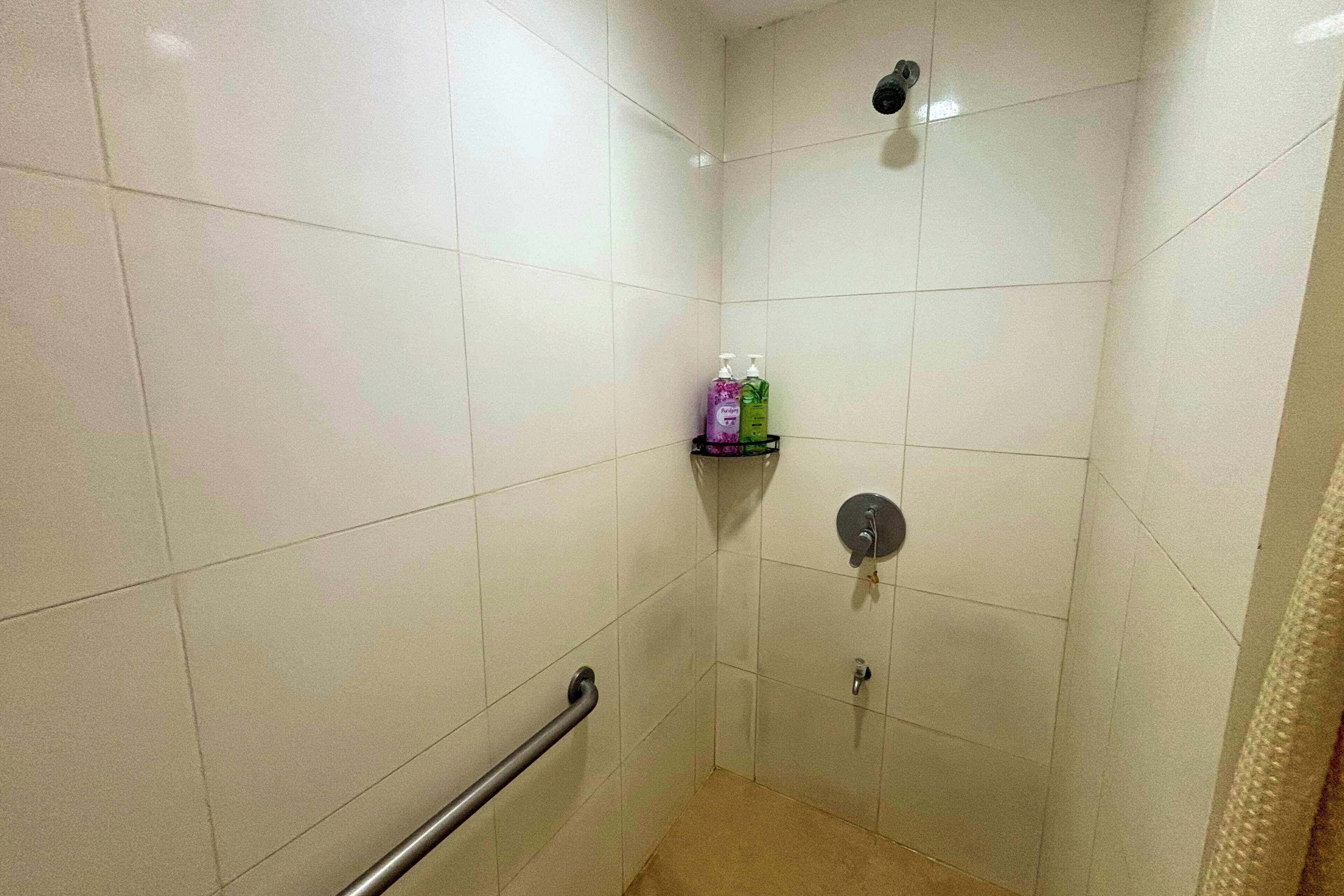 A small, tiled shower area with a handrail, a shelf with toiletries, and a showerhead.