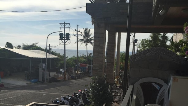 Un balcón con sillas y mesas de metal blanco, con vista a una calle donde se ven motocicletas estacionadas y un edificio con tejado inclinado.