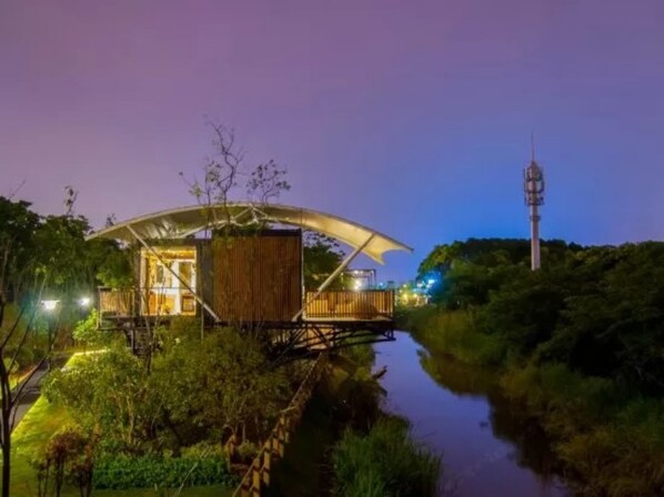 A modern, elevated wooden structure with a large canopy, situated by a calm river, surrounded by lush greenery. - Lin Jia Camping Site Shanghai (Shanghai)