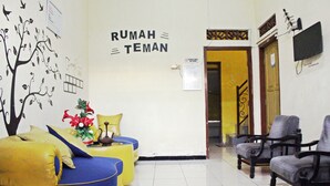 A reception area with a wall sign reading "RUMAH TEMAN" and a tree mural. There are yellow and blue seating areas with a vase of flowers.