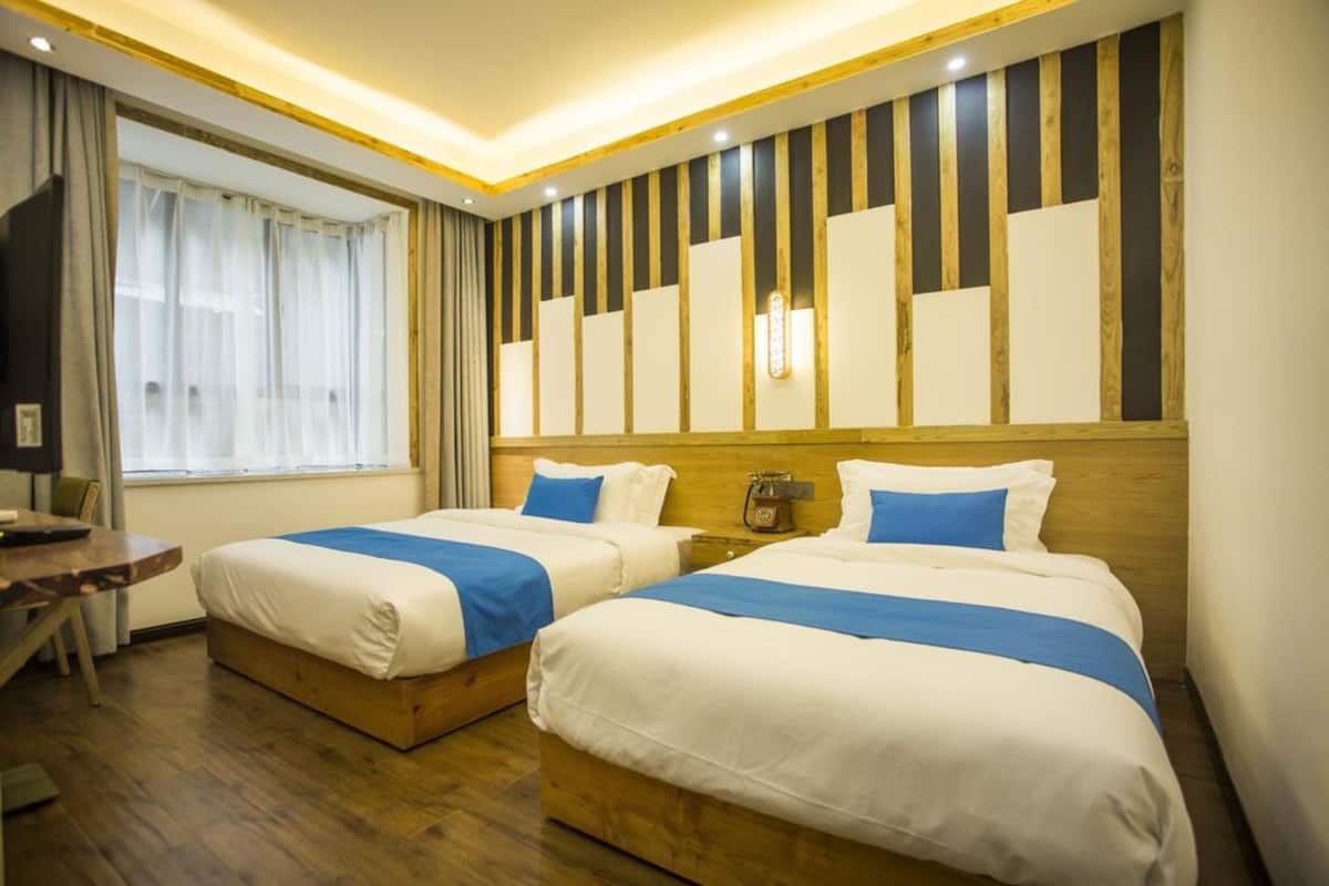A hotel room with two beds, a wooden headboard, a desk, and a large window with curtains.