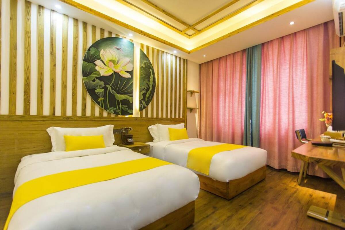 A hotel room with two beds, wooden headboards, a large floral artwork on the wall, a desk with a chair, and a window with pink curtains.