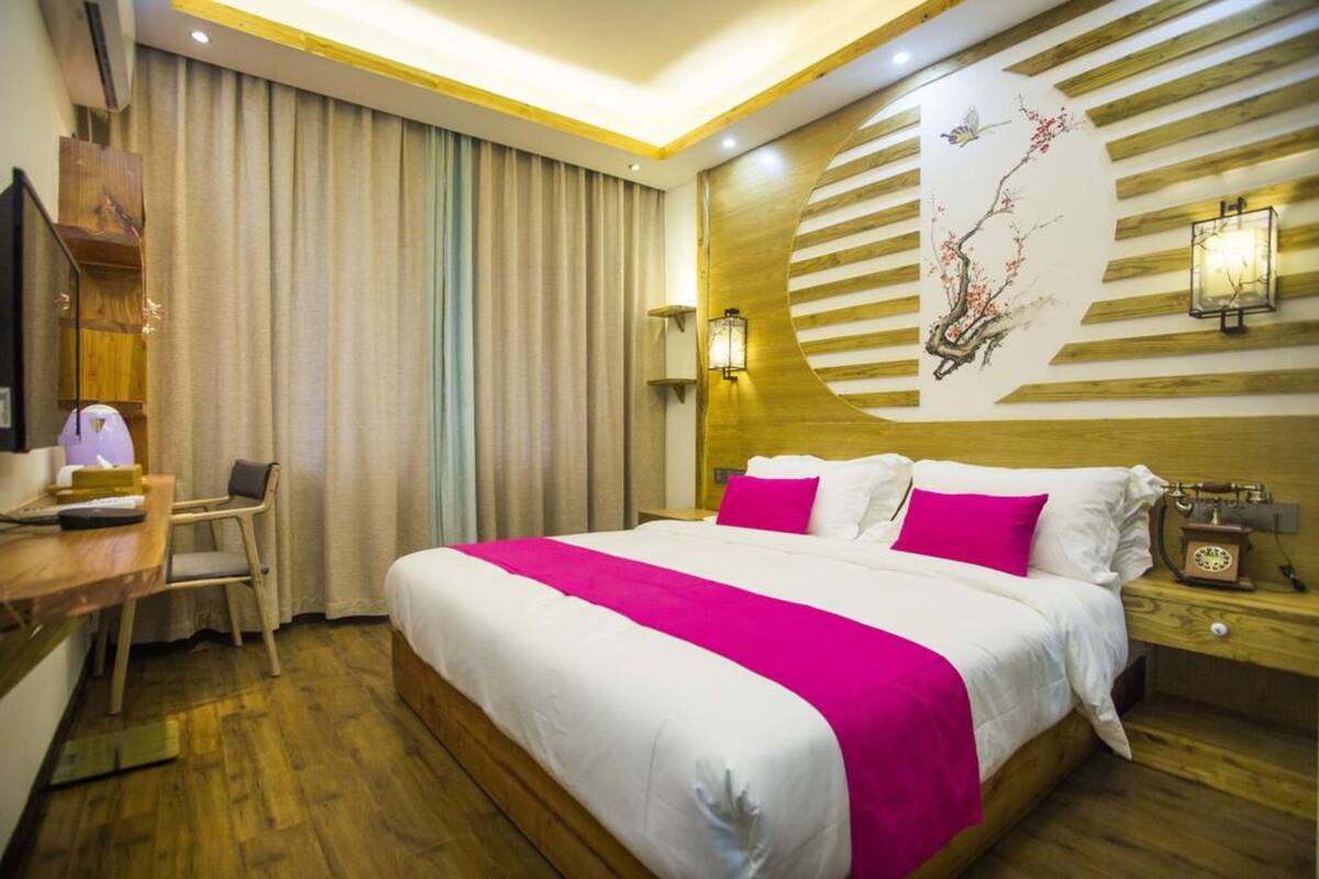 A hotel room with a large bed, a desk with a chair, a nightstand with a lamp, and a wall with a decorative panel.