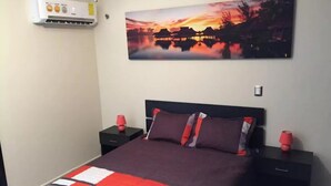 A bedroom with a bed, two bedside tables, a nightstand, a wall-mounted air conditioner, and a painting of a sunset over water.