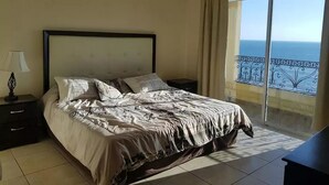 A bedroom with a bed, a nightstand, a lamp, and a view of the ocean through a large window. - Condominio Malecon (Mazatlán)