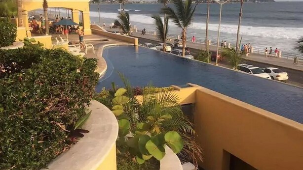 A pool area with a curved design, surrounded by palm trees and a view of the ocean. - Condominio Malecon (Mazatlán)