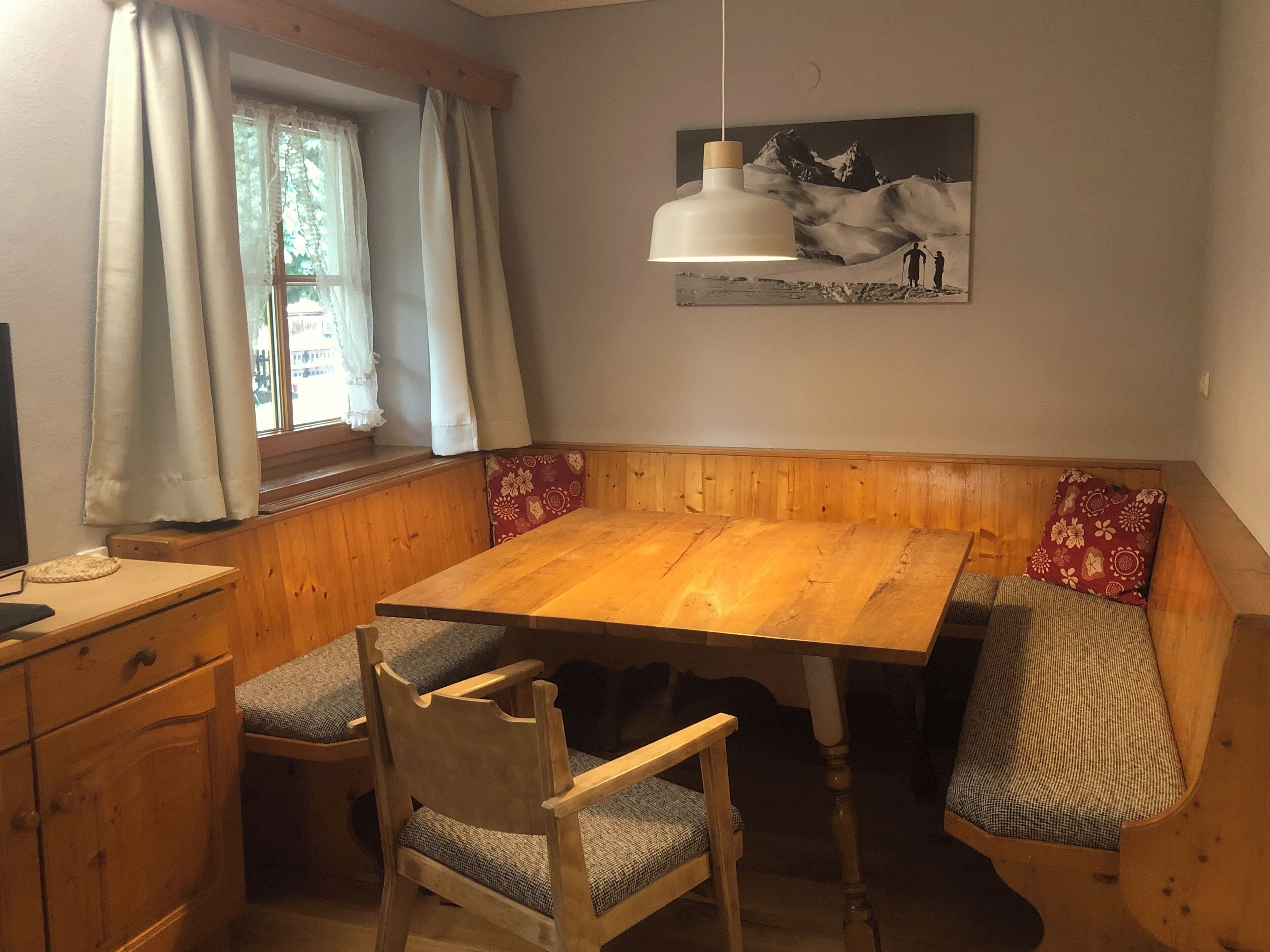 A dining area with a wooden table and bench seating, a window with curtains, and a hanging light fixture.