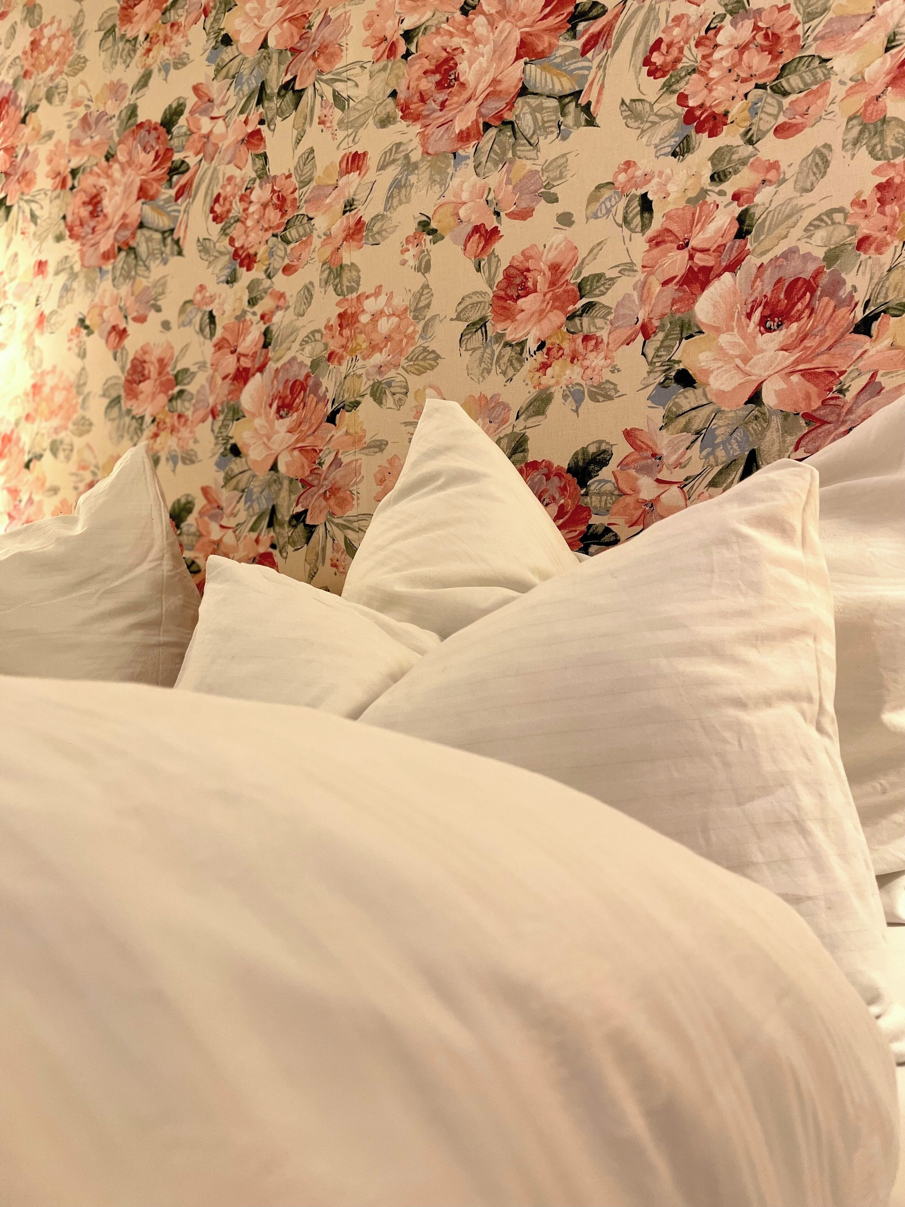 A bed with white bedding and a floral wallpaper background.