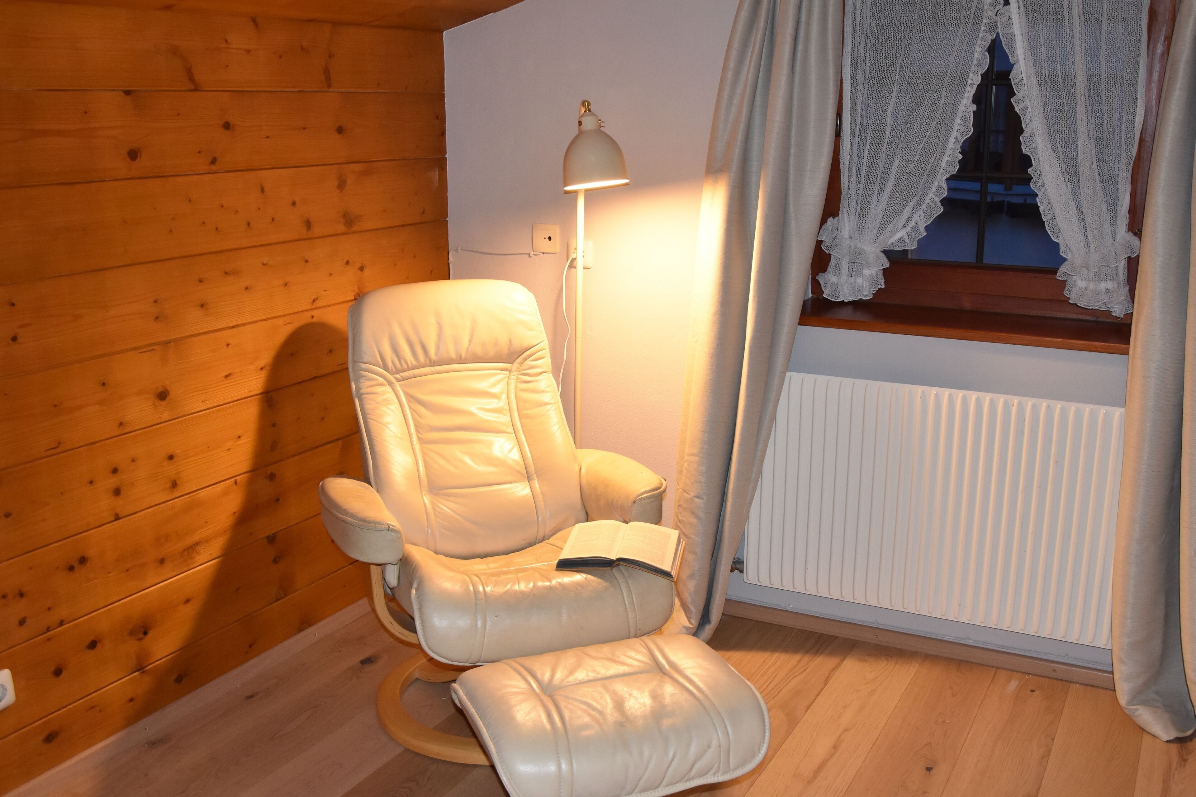 A room with a wooden wall, a white radiator, a white chair with a book, and a floor lamp.