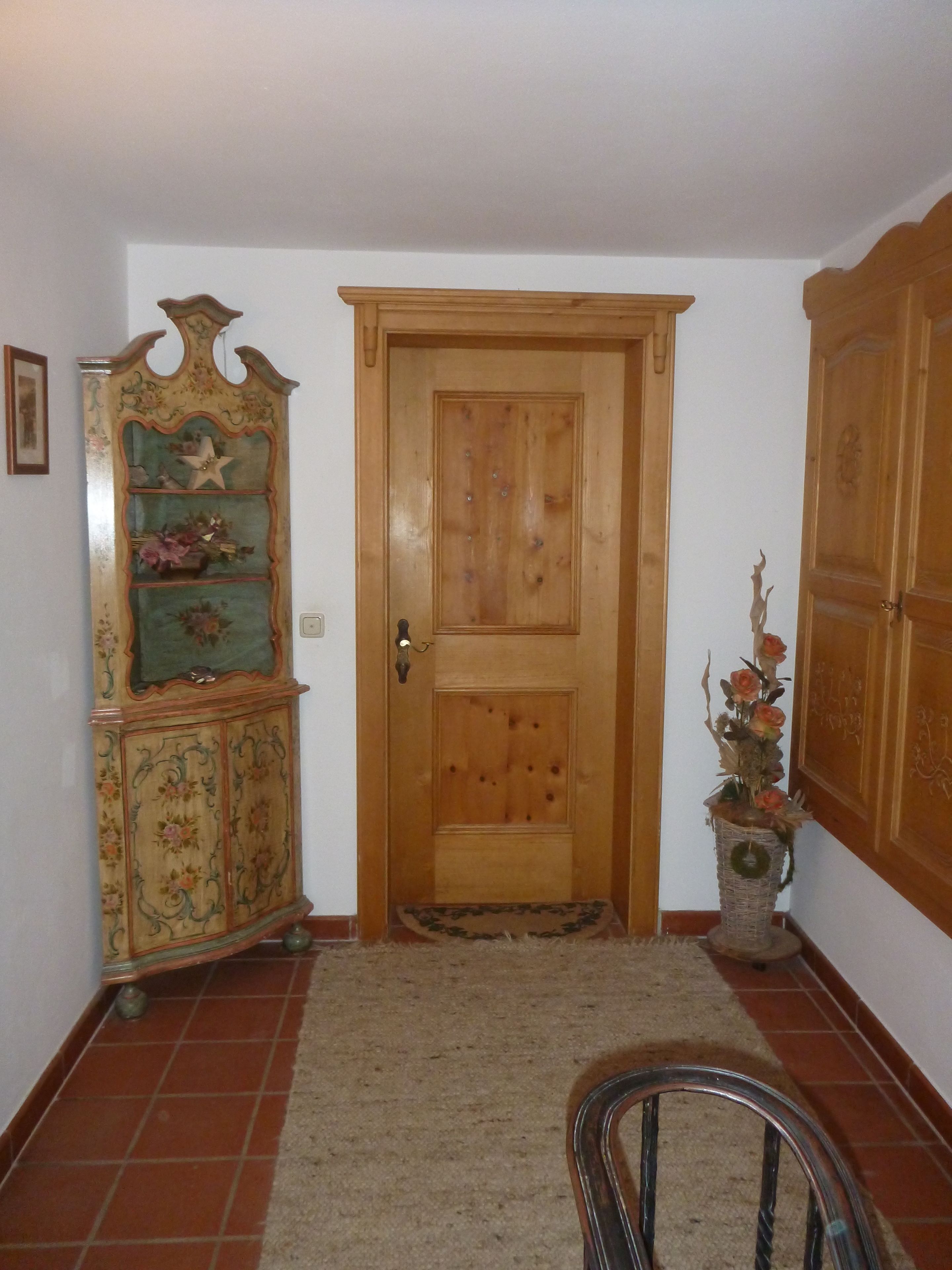 Eine Holztür mit einer kleinen Matte davor, ein Holzschrank mit floralem Muster links daneben und eine Korbvase mit getrockneten Blumen zur rechten Seite.