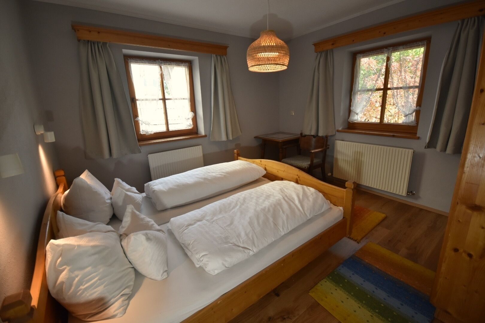 Ein Schlafzimmer mit zwei Einzelbetten, Holz-Kopfteilen, einem Holztisch, zwei mit Vorhängen versehenen Fenstern und einer Pendelleuchte.