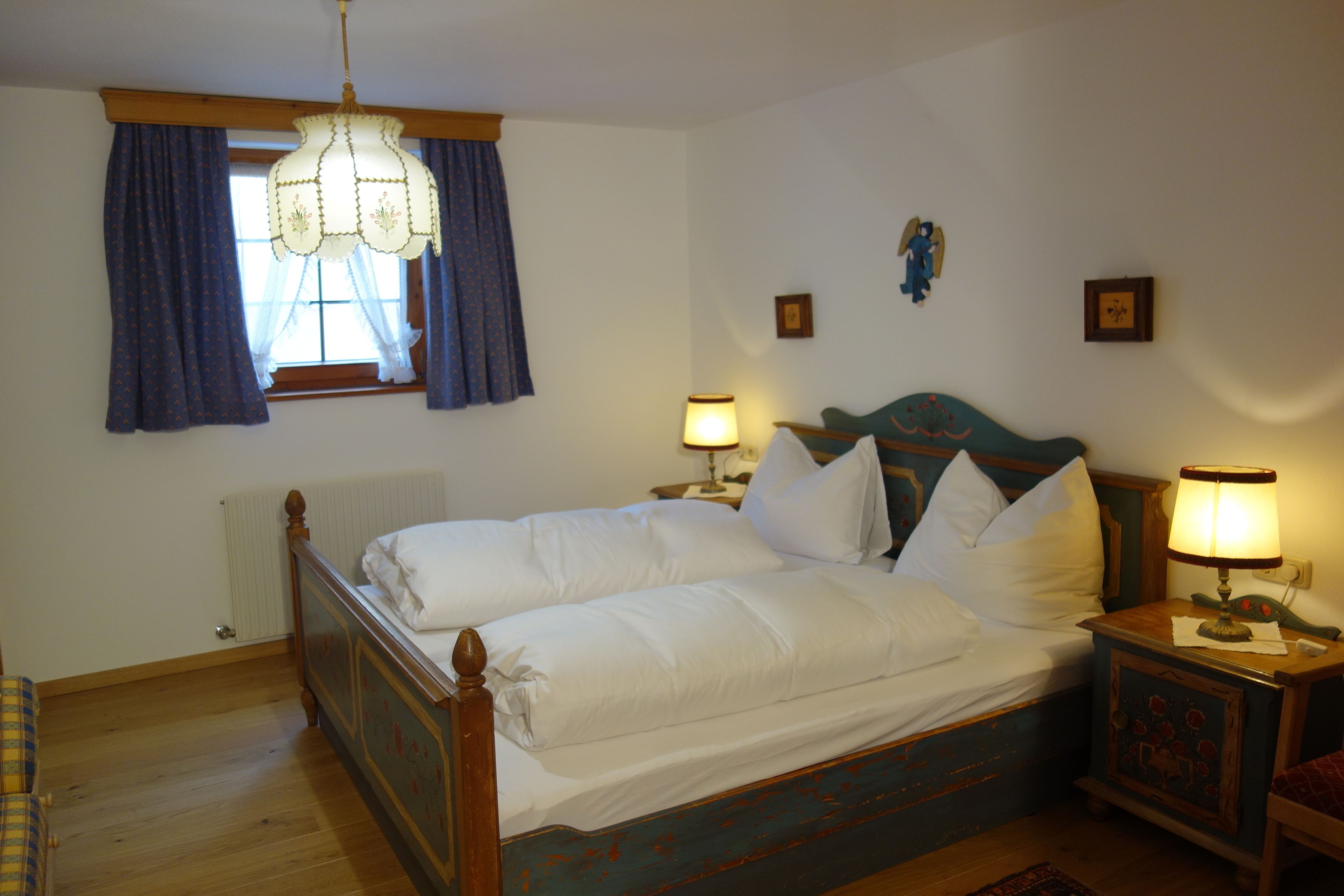 A bedroom with a wooden bed, two bedside tables with lamps, a window with curtains, and a radiator.