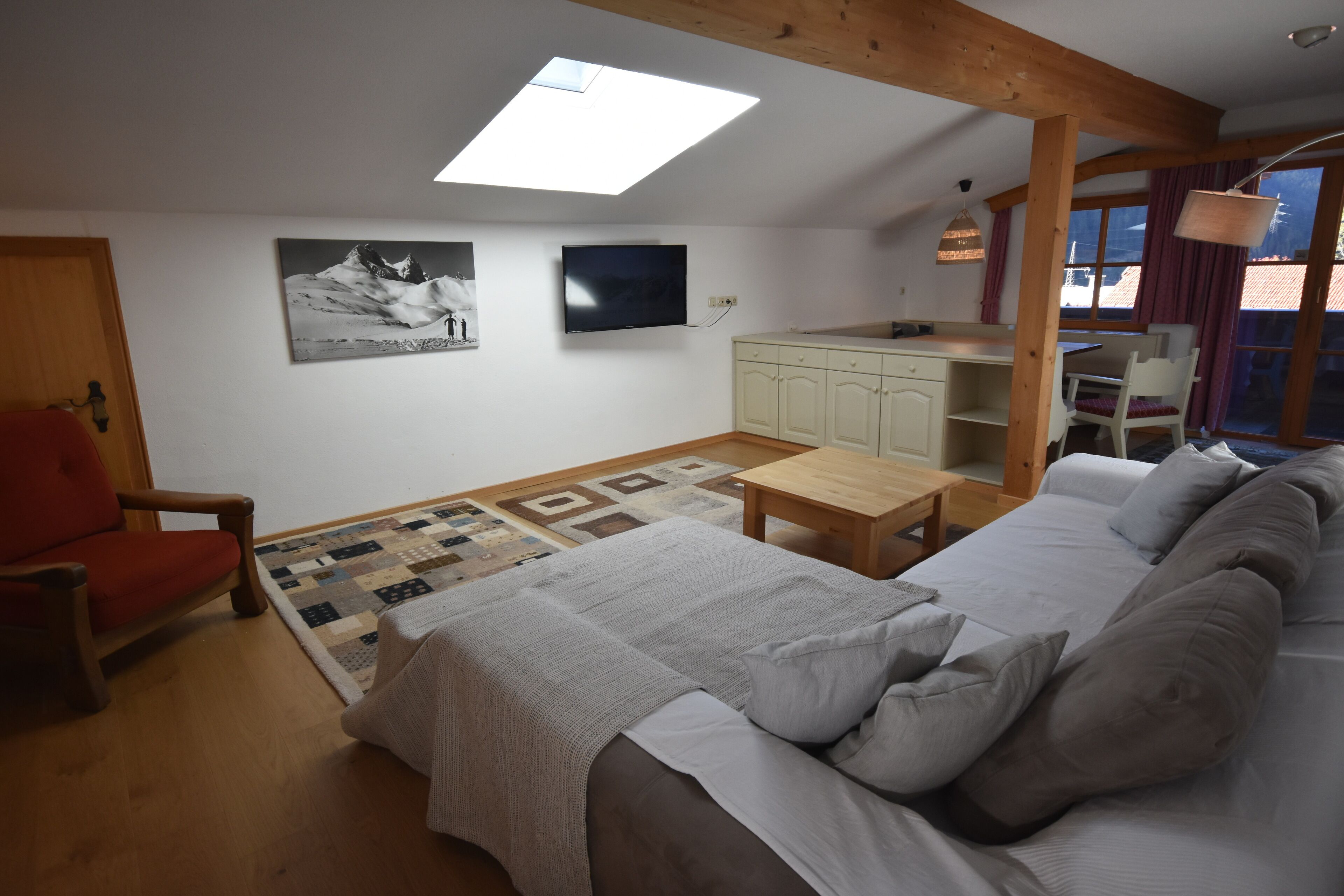 A cozy room with a bed, a red chair, a wooden table, a TV, and a kitchen area.
