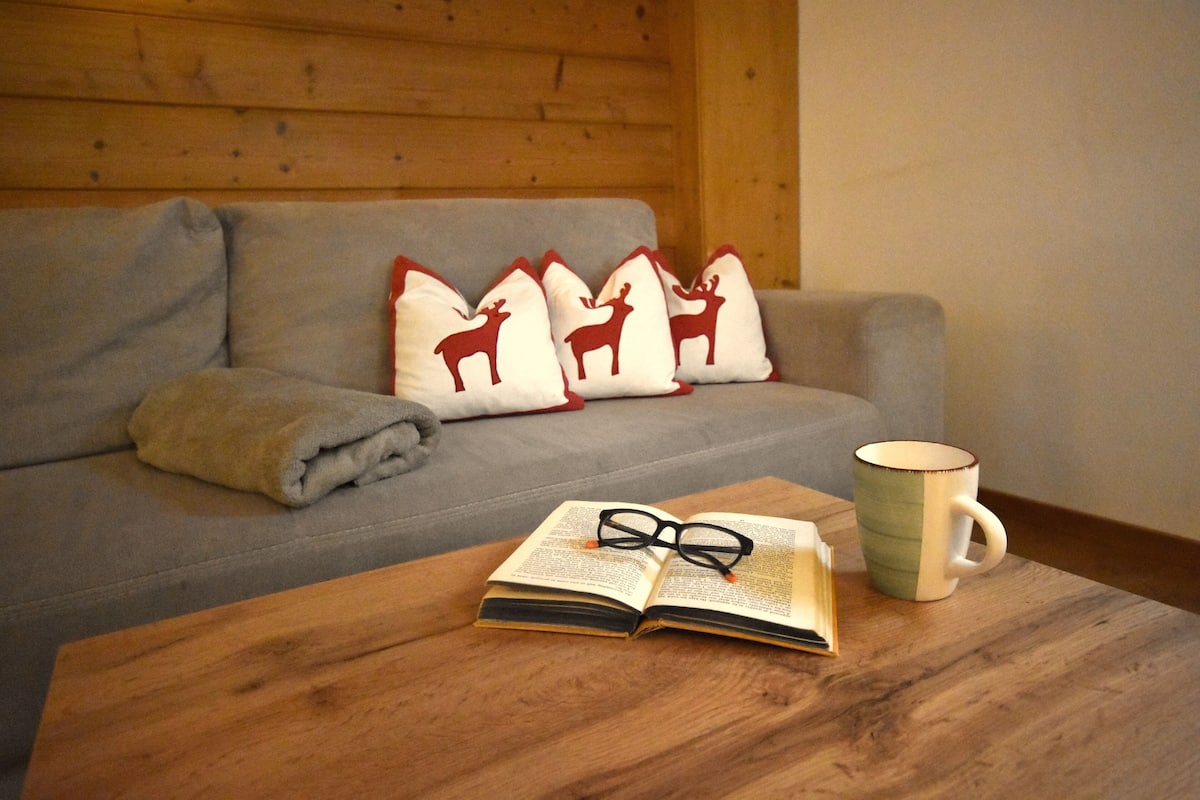 A cozy room with a wooden table, a grey sofa with decorative pillows, an open book, and a mug.