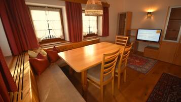 A cozy room with a wooden dining table, benches, and chairs. There is a television on a stand, a window with curtains, and a small cabinet.