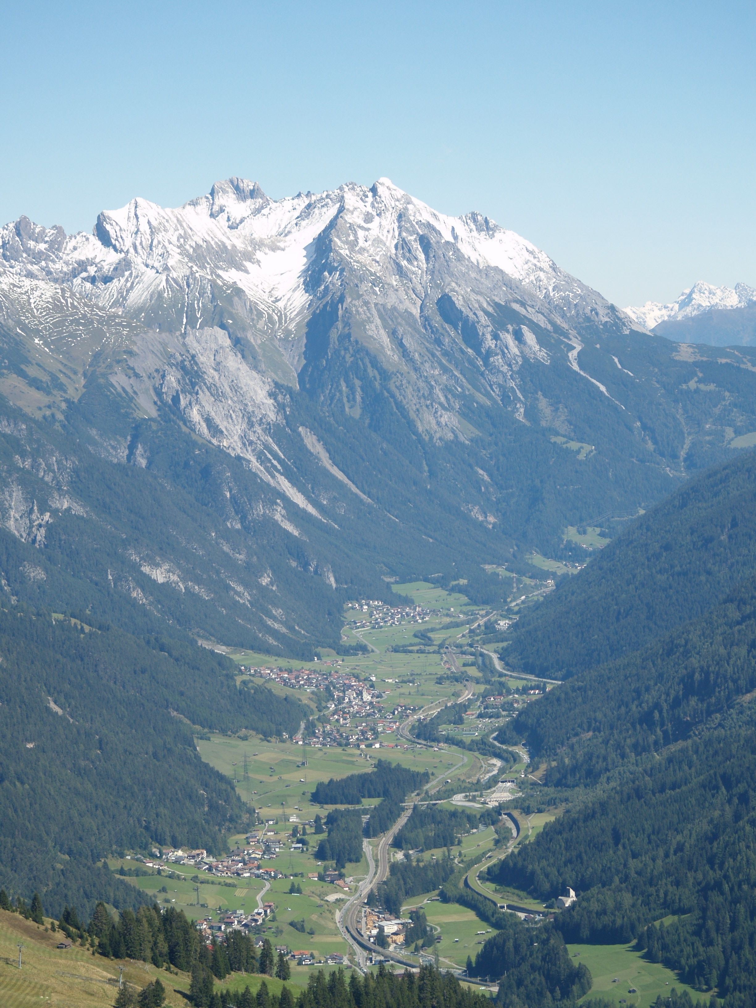 A mountainous landscape with a valley, a winding road, and a small town with buildings and greenery.