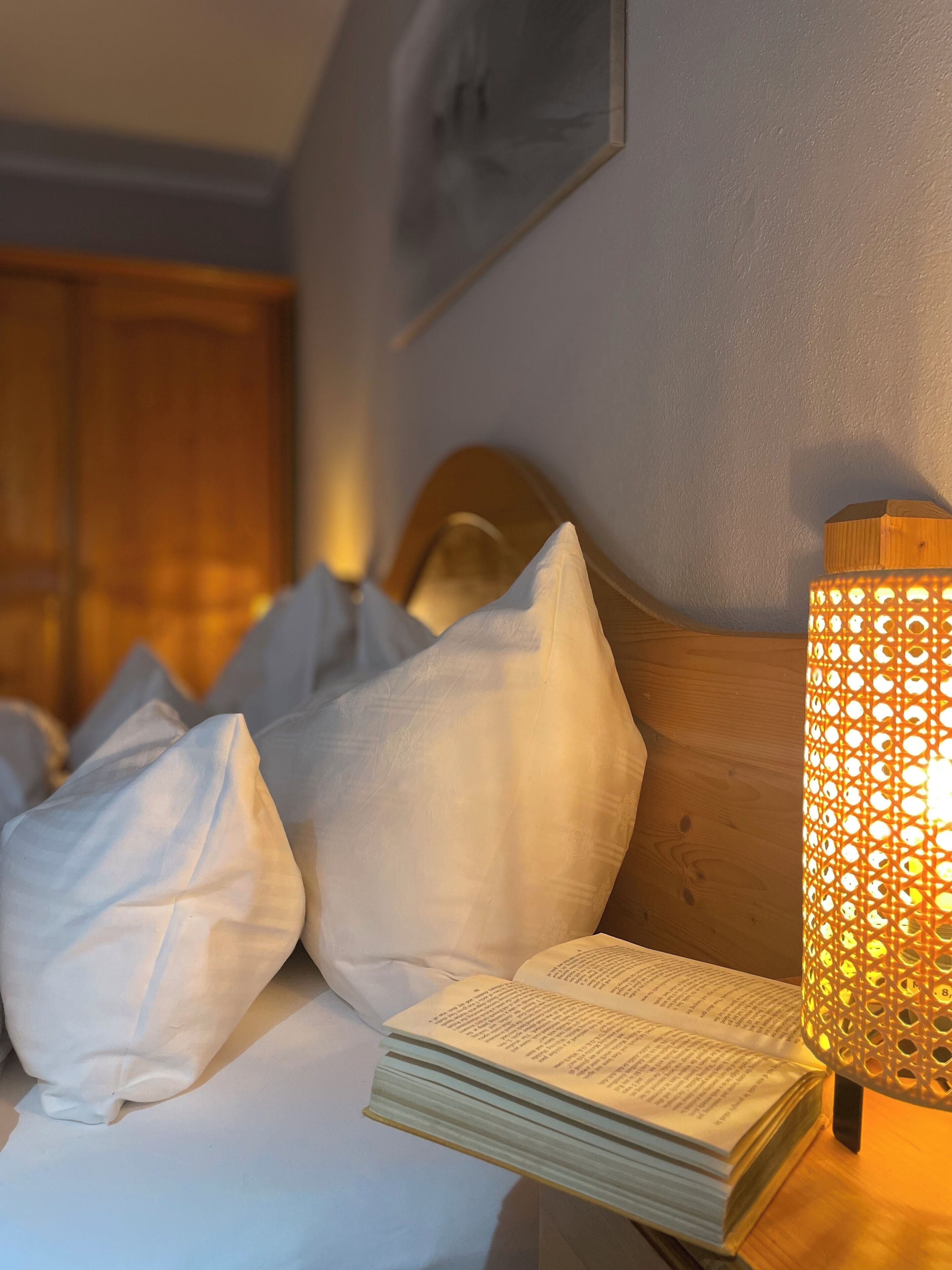Una cama bien tendida con almohadas y un libro abierto, cabecera de madera y un cuadro colgado en la pared.