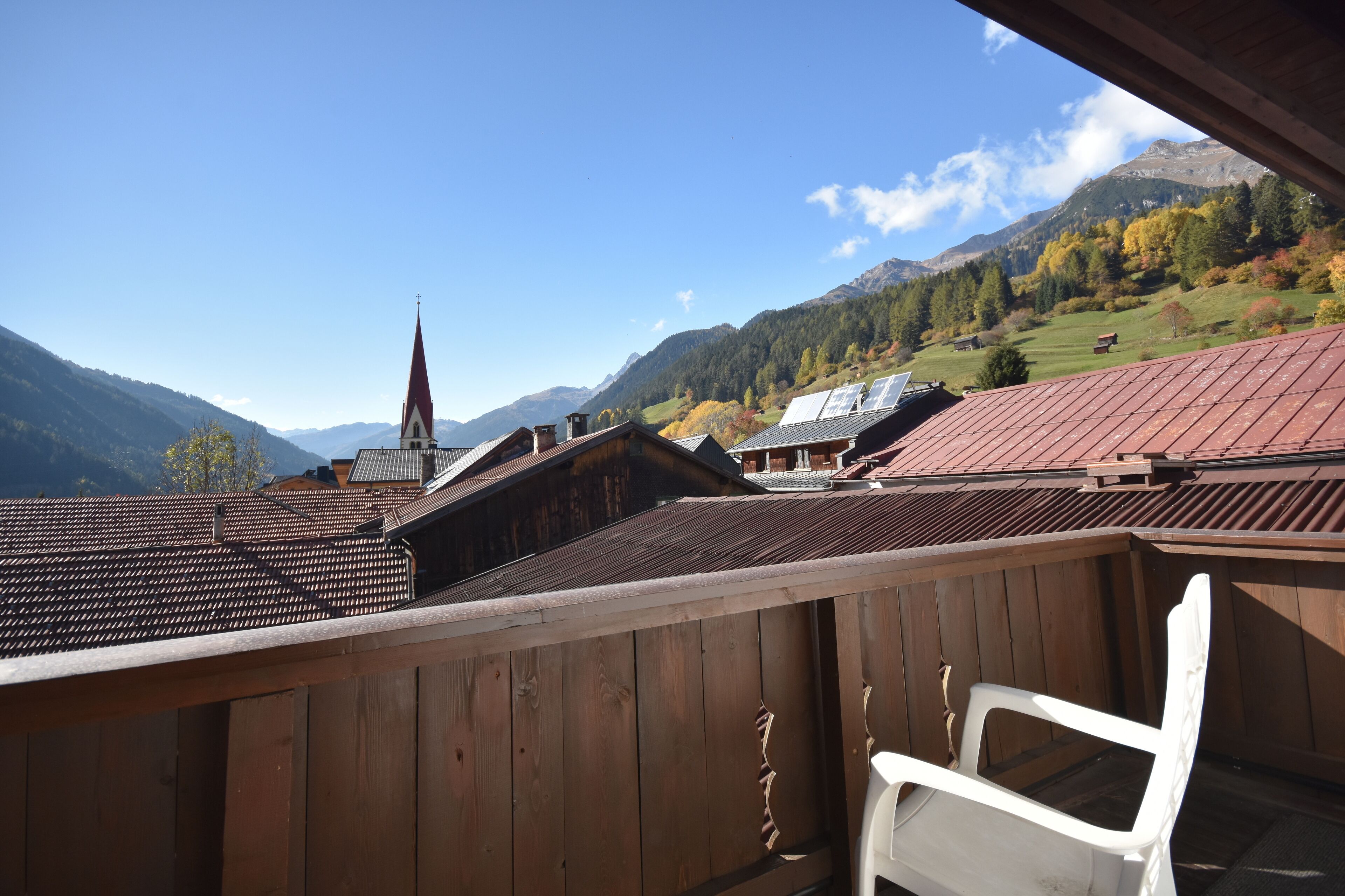 A wooden balcony with a white chair, overlooking a village with traditional buildings and a church spire, set against a backdrop of mountains and autumn foliage.