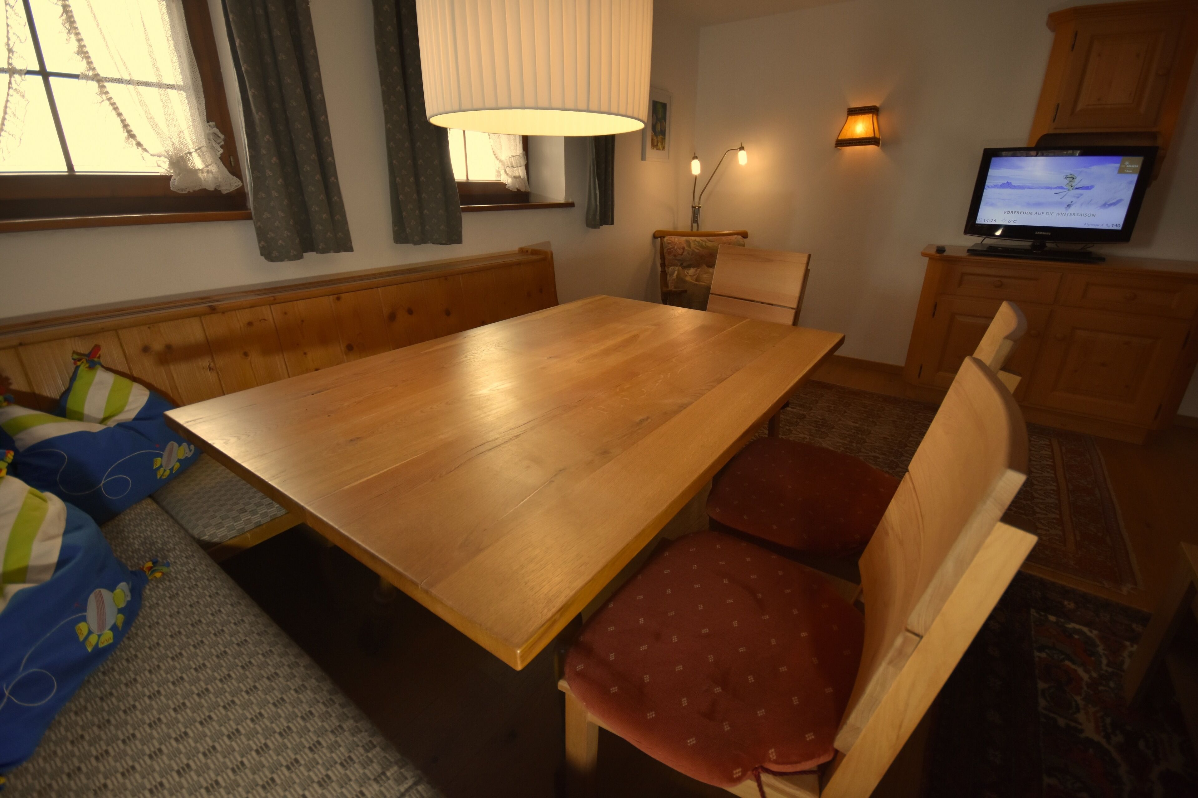 Una habitación acogedora con una mesa de madera, un banco con cojines rojos, un televisor y una lámpara.