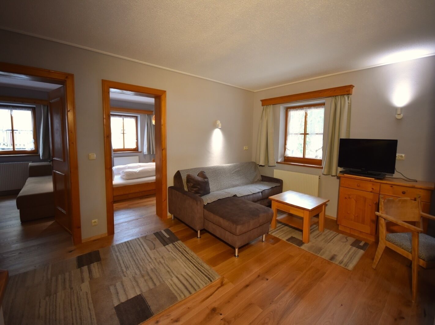 A compact living space with a sofa, a small table, a TV, and a wooden cabinet. There are two windows with curtains, and a door leading to another room.