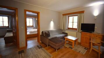 A compact living space with a sofa, a small table, a TV, and a wooden cabinet. There are two windows with curtains, and a door leading to another room.