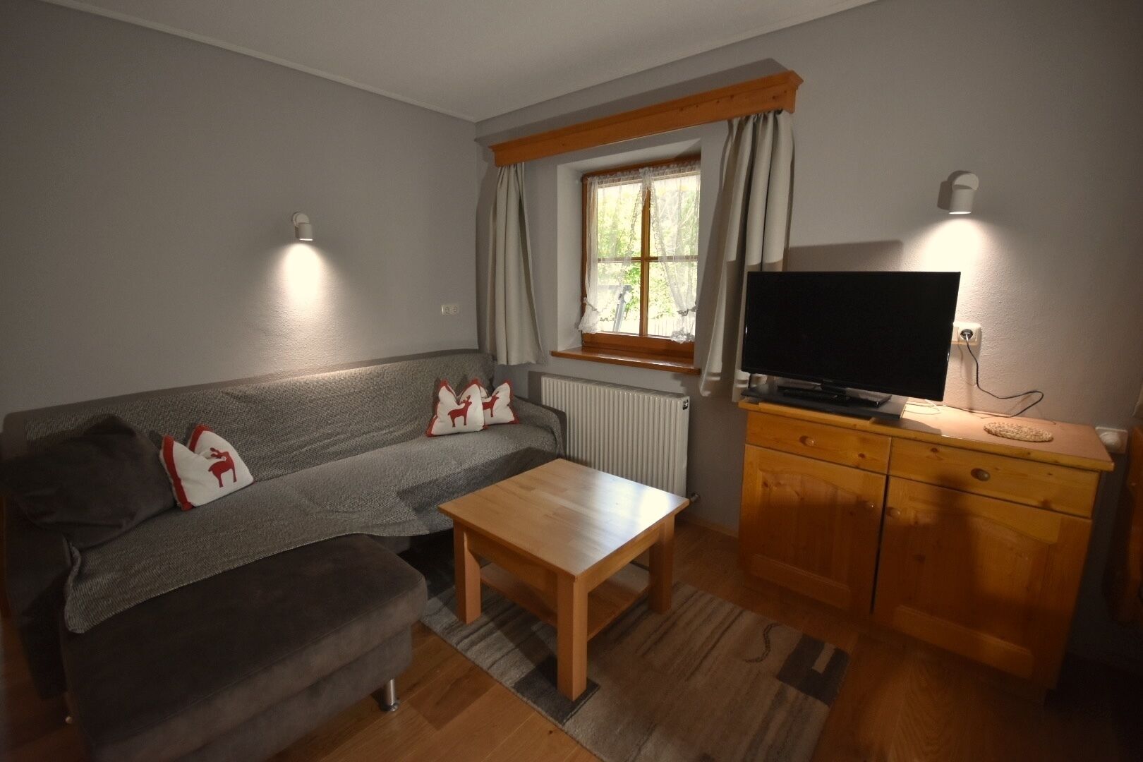 Una sala de estar acogedora con un sofá gris, una mesa de centro de madera, un televisor sobre un mueble de madera y una ventana con cortinas.