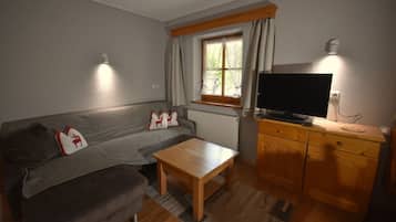 Una sala de estar acogedora con un sofá gris, una mesa de centro de madera, un televisor sobre un mueble de madera y una ventana con cortinas.