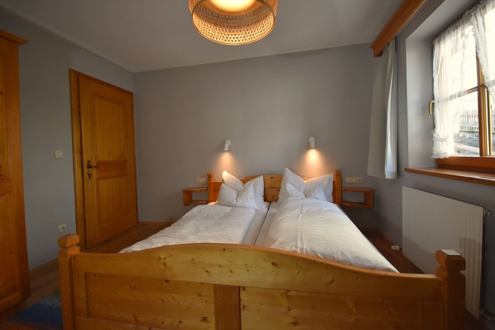 Ein Doppelbett aus Holz mit zwei Nachttischen, ein Holzschrank, ein Fenster mit Vorhängen und eine Deckenleuchte.