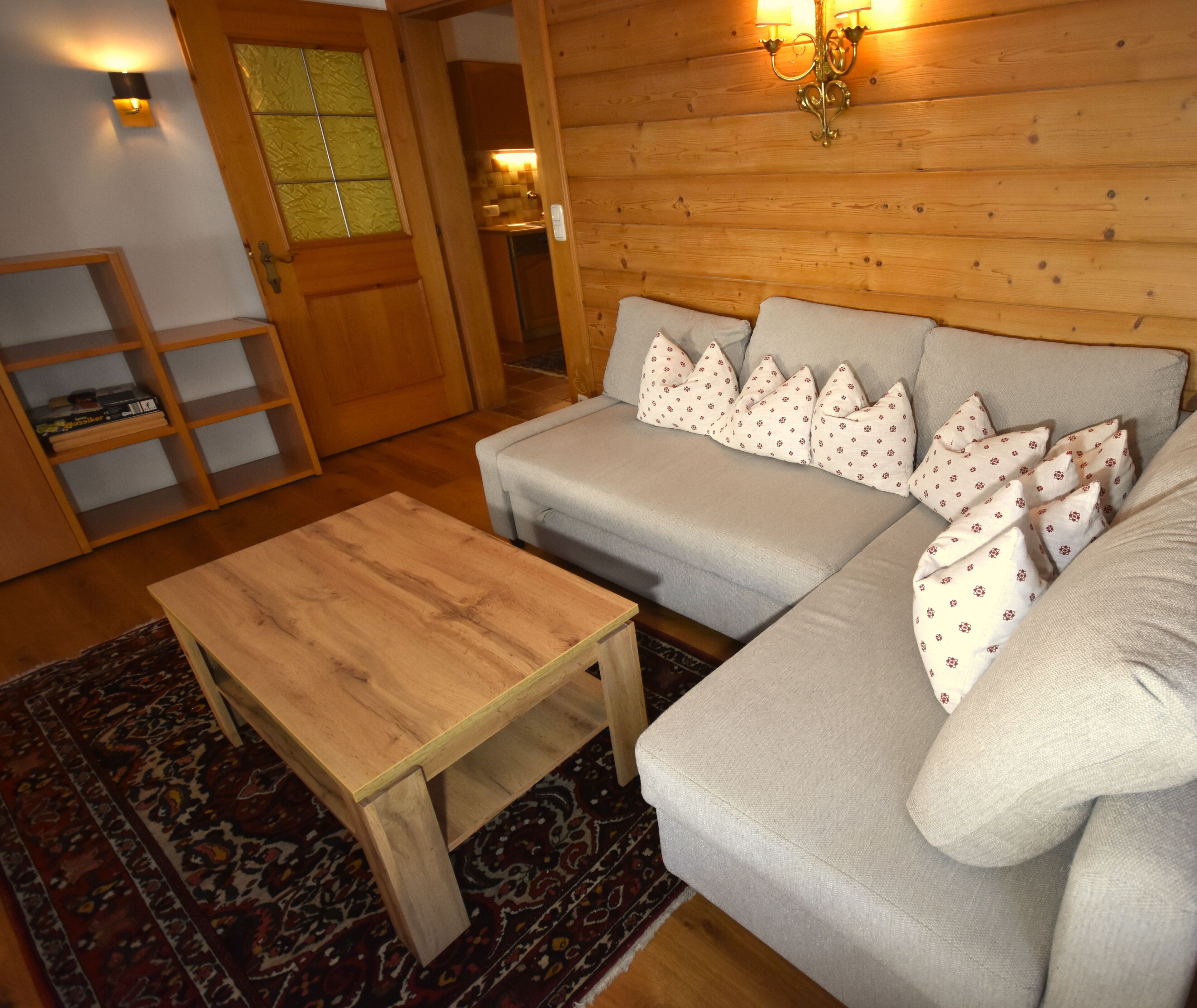 Ein gemütliches Wohnzimmer mit einem Holztisch, einer grauen Couch mit gemusterten Kissen und einem Bücherregal aus Holz.