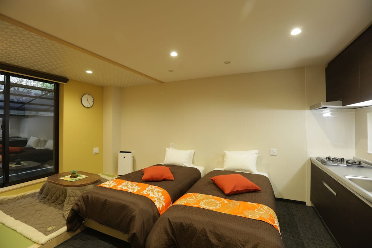 Habitación de hotel con dos camas, una mesita, una ventana, un reloj y un kitchenette.