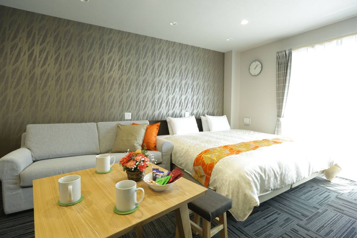 Habitación de hotel con sofá, cama, mesa con tazas y frutero, y ventana con cortinas.