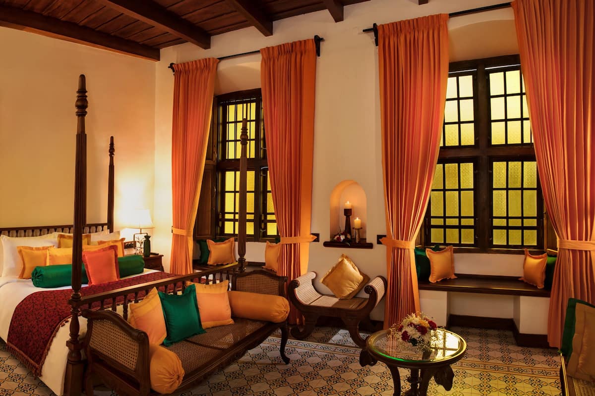 A traditional room with a four-poster bed, a sitting area with a bench, and two windows with curtains.