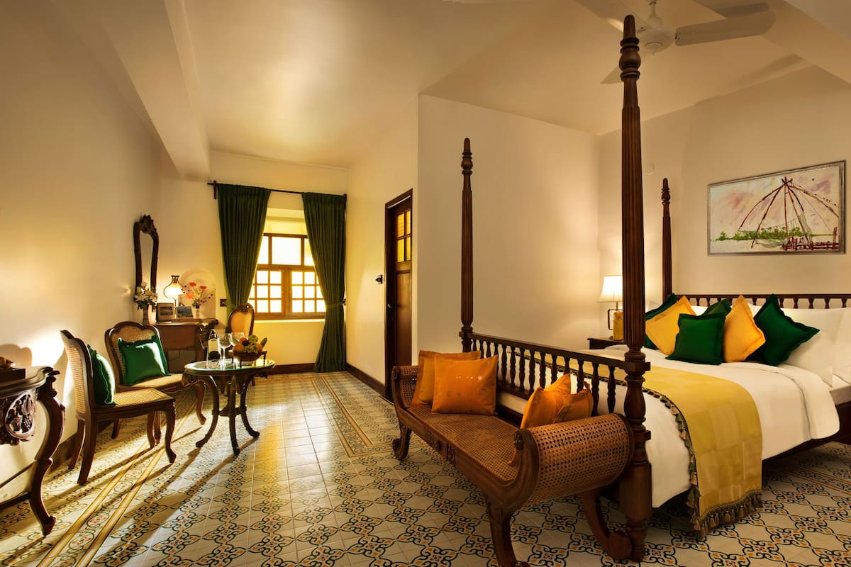 A four-poster bed with a canopy, a sitting area with a chair and table, a small desk with a lamp, and a patterned floor.