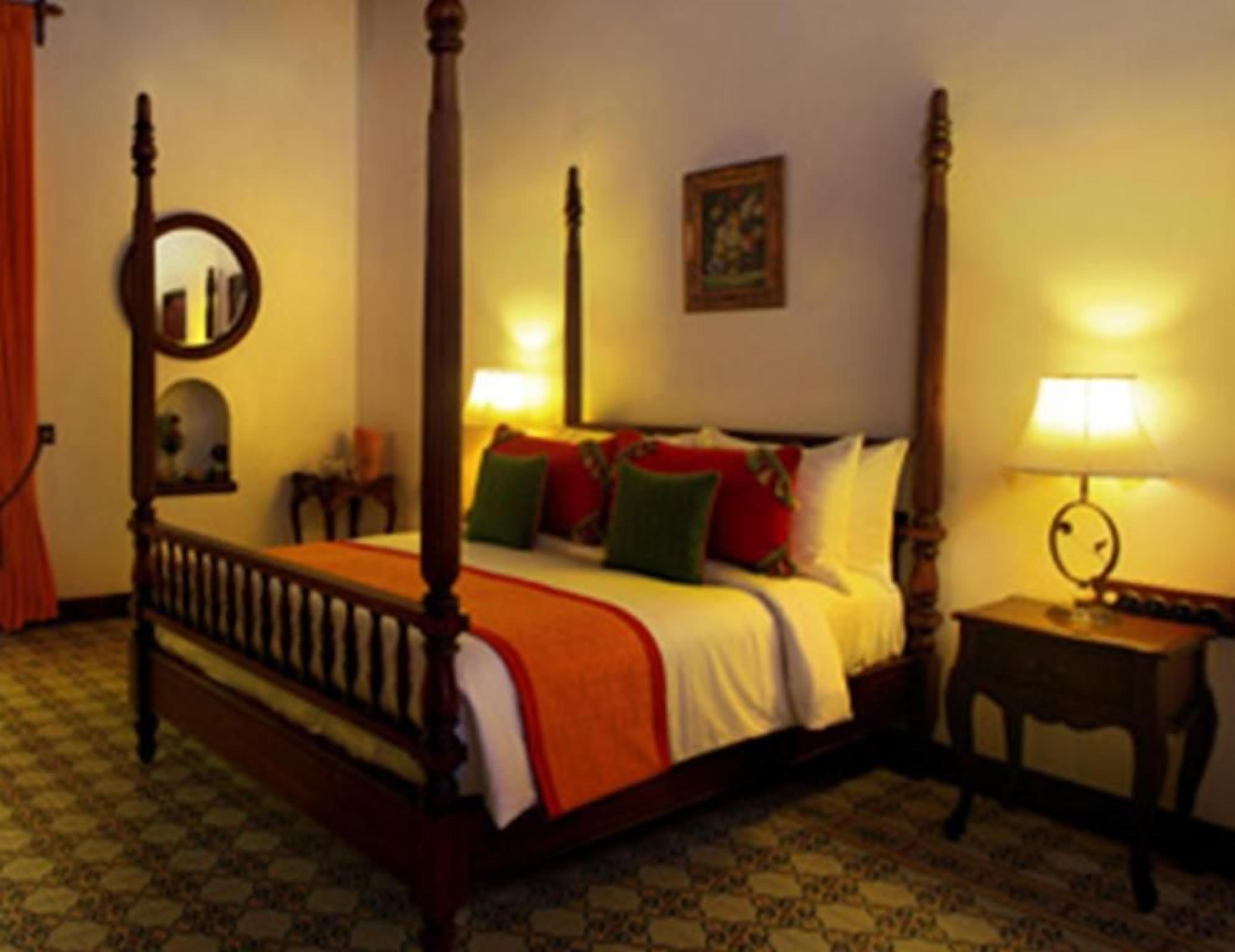 A four-poster bed with a wooden frame and a white bedspread, a bedside table with a lamp, and a patterned floor.