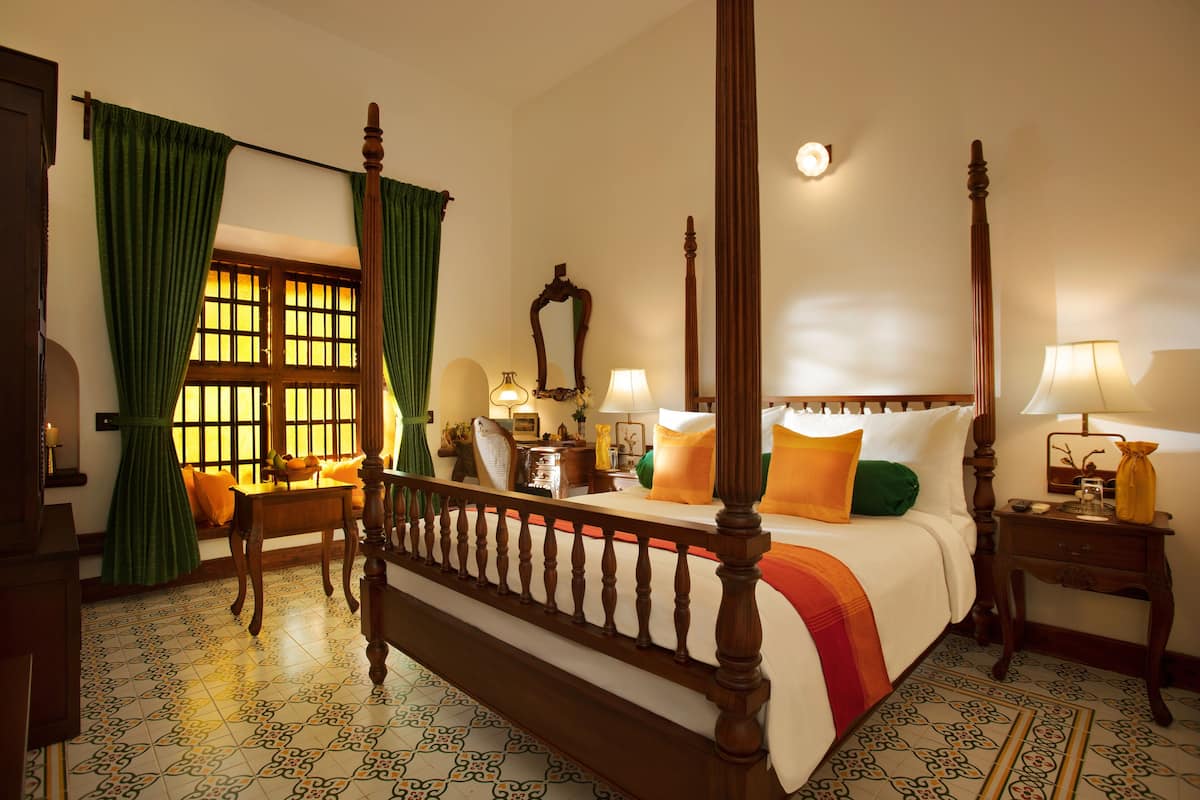 A four-poster bed with a wooden headboard, a nightstand with a lamp, and a window with green curtains.