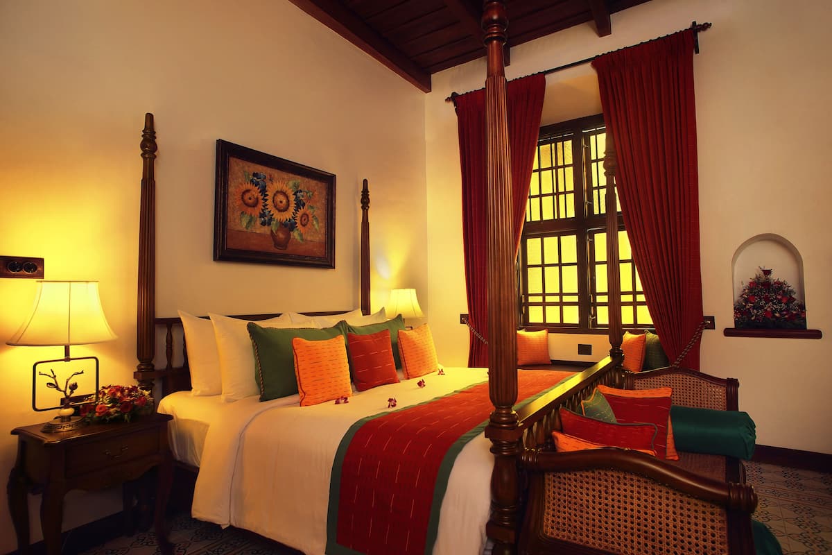 A hotel room with a four-poster bed, a nightstand with a lamp, a painting on the wall, and a window with curtains.