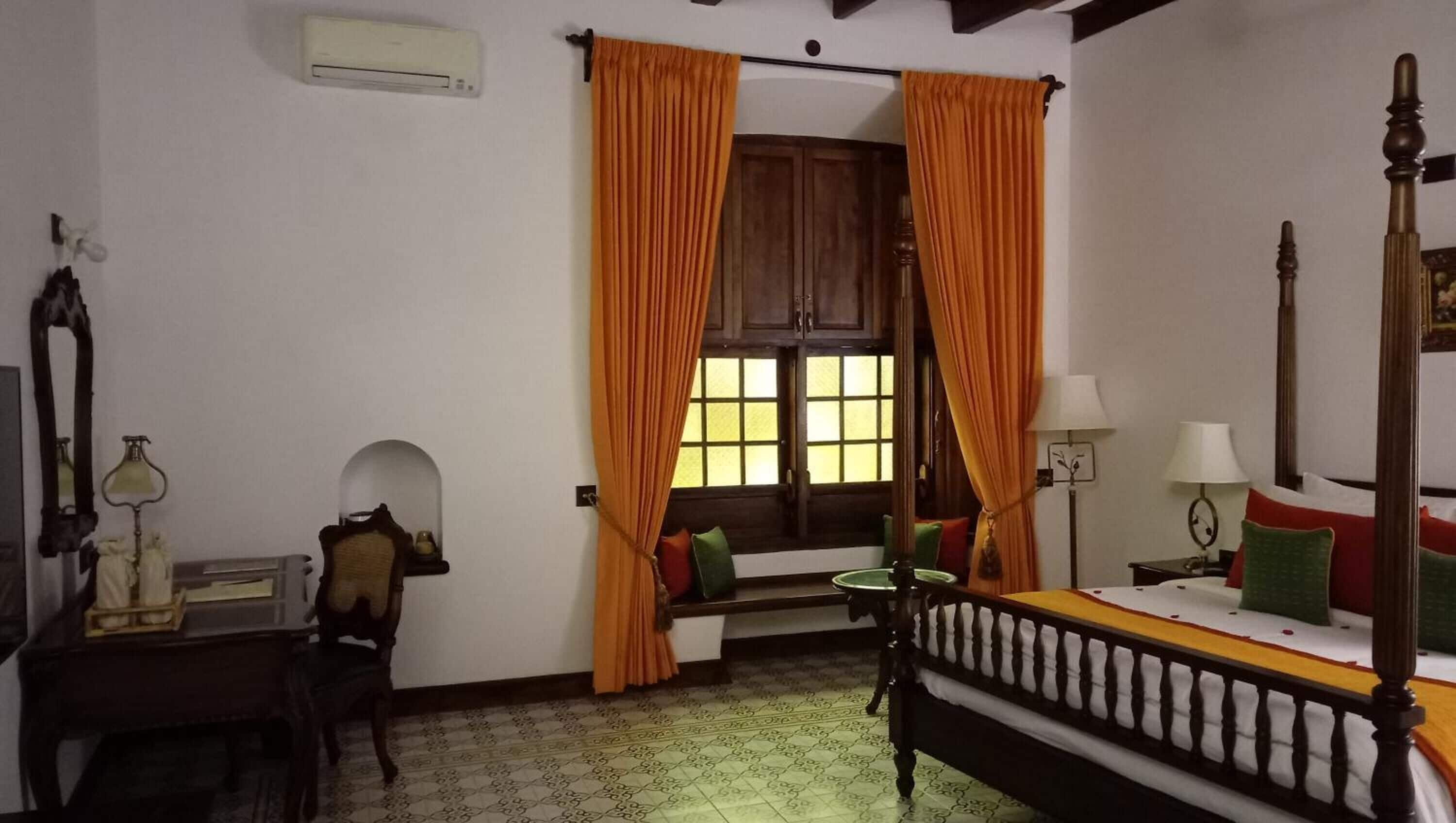 Ein Zimmer mit Bett, Sessel, Schreibtisch, einem Fenster mit Vorhängen und einem Spiegel.