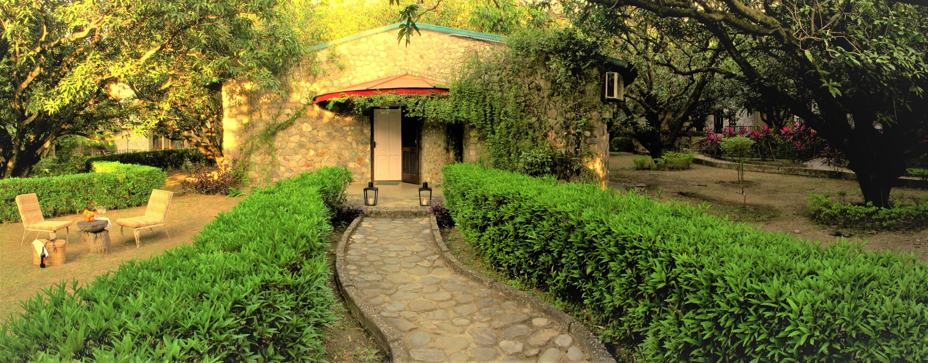 A stone pathway leading to a rustic stone building with a red roof, surrounded by lush greenery and a small seating area with wicker chairs and a table.