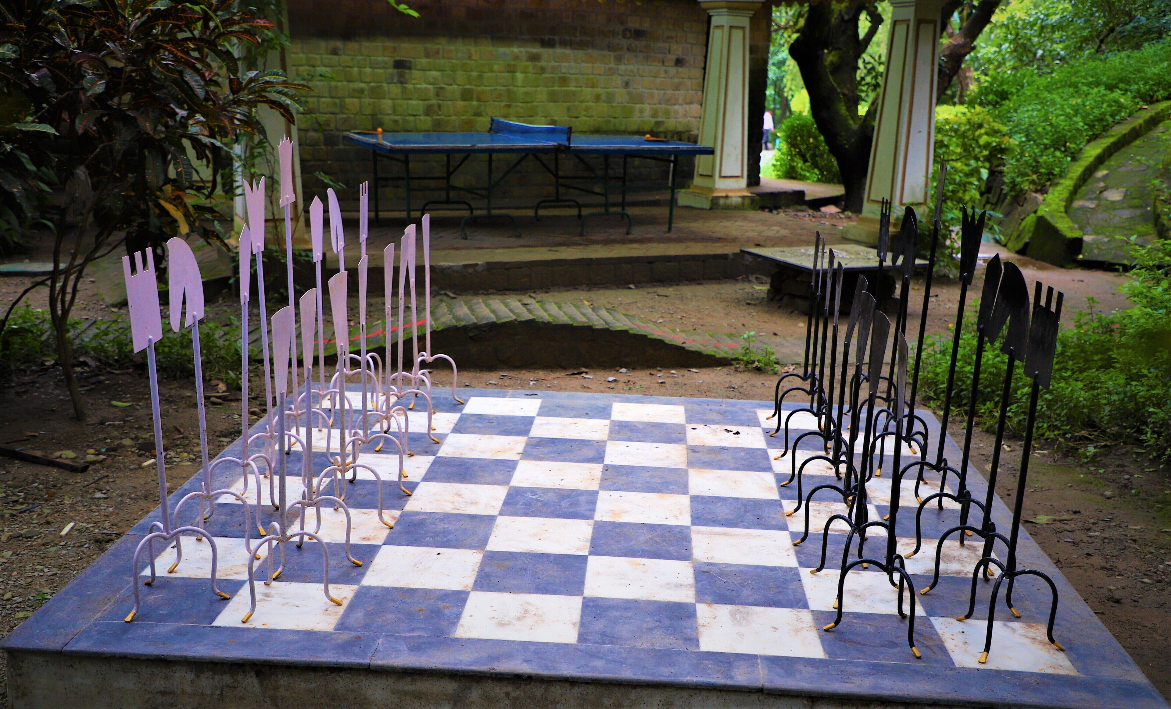 A checkered outdoor table with metal stands, surrounded by greenery and a brick wall.