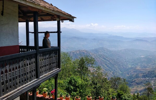 Un balcón con una persona de pie, con vista a un paisaje montañoso y campos en terrazas.