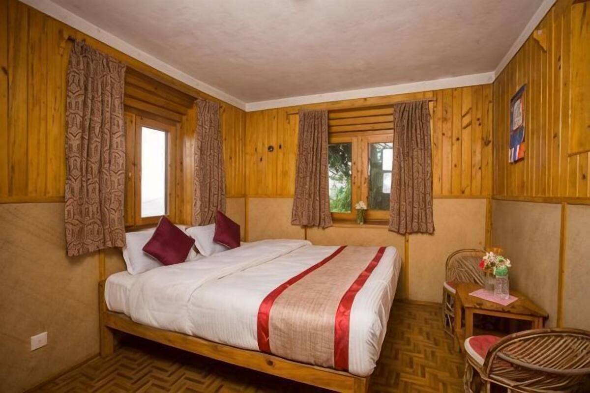 Habitación con paneles de madera, una cama, cortinas, una mesita con un florero y una silla.