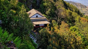 Una casa tradicional con techo inclinado, rodeada de abundante vegetación y montañas al fondo.