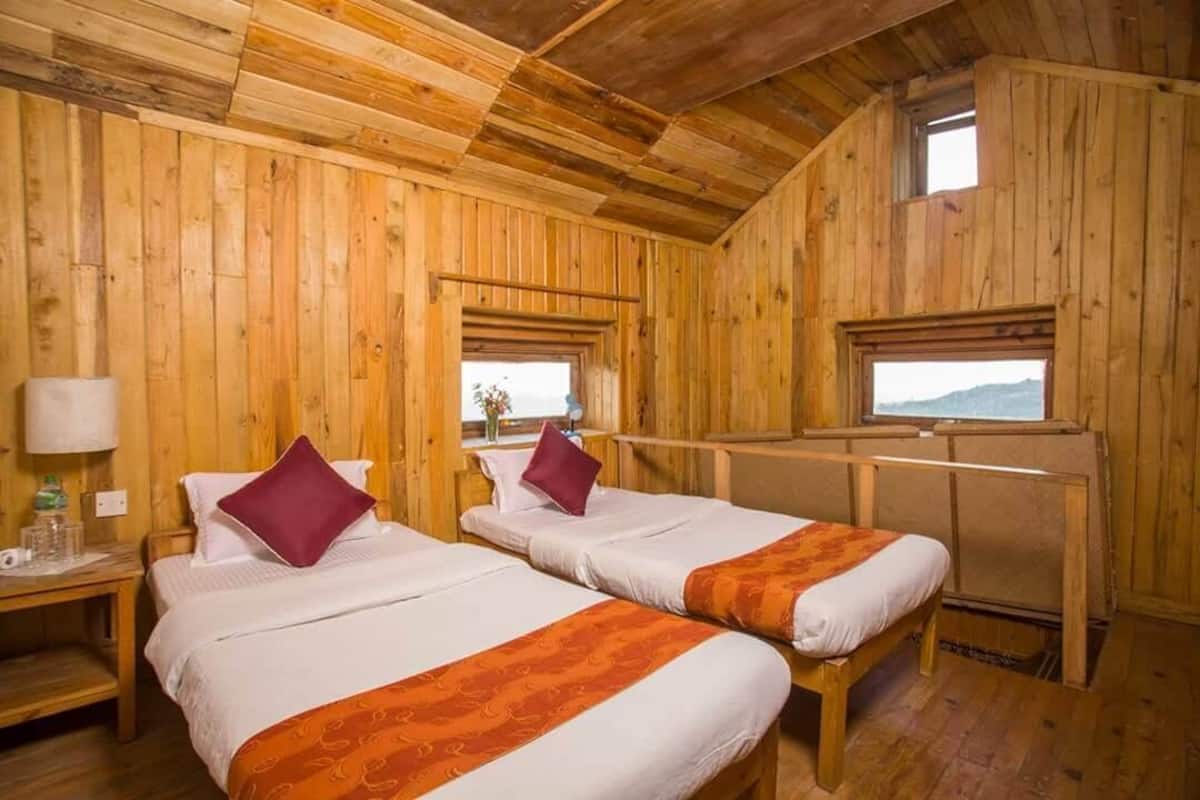Habitación de cabaña de madera con dos camas, cada una con almohada y manta. Hay una ventana en la pared derecha y una pequeña mesa con una lámpara y una botella a la izquierda.